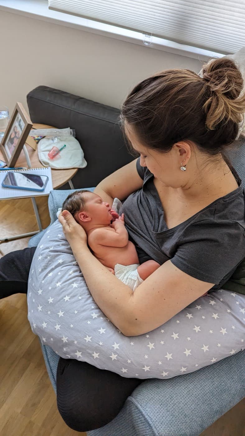 Mãe amamentando o recém-nascido em casa, apoiada por uma almofada de amamentação